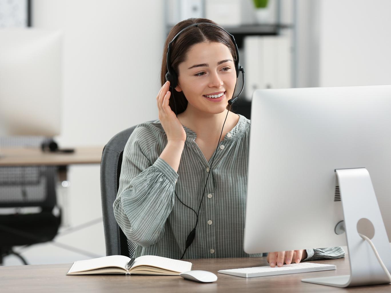 Eine lächelnde Frau mit Headset sitzt vor einem Computer und hält sich die Hand ans Ohr.