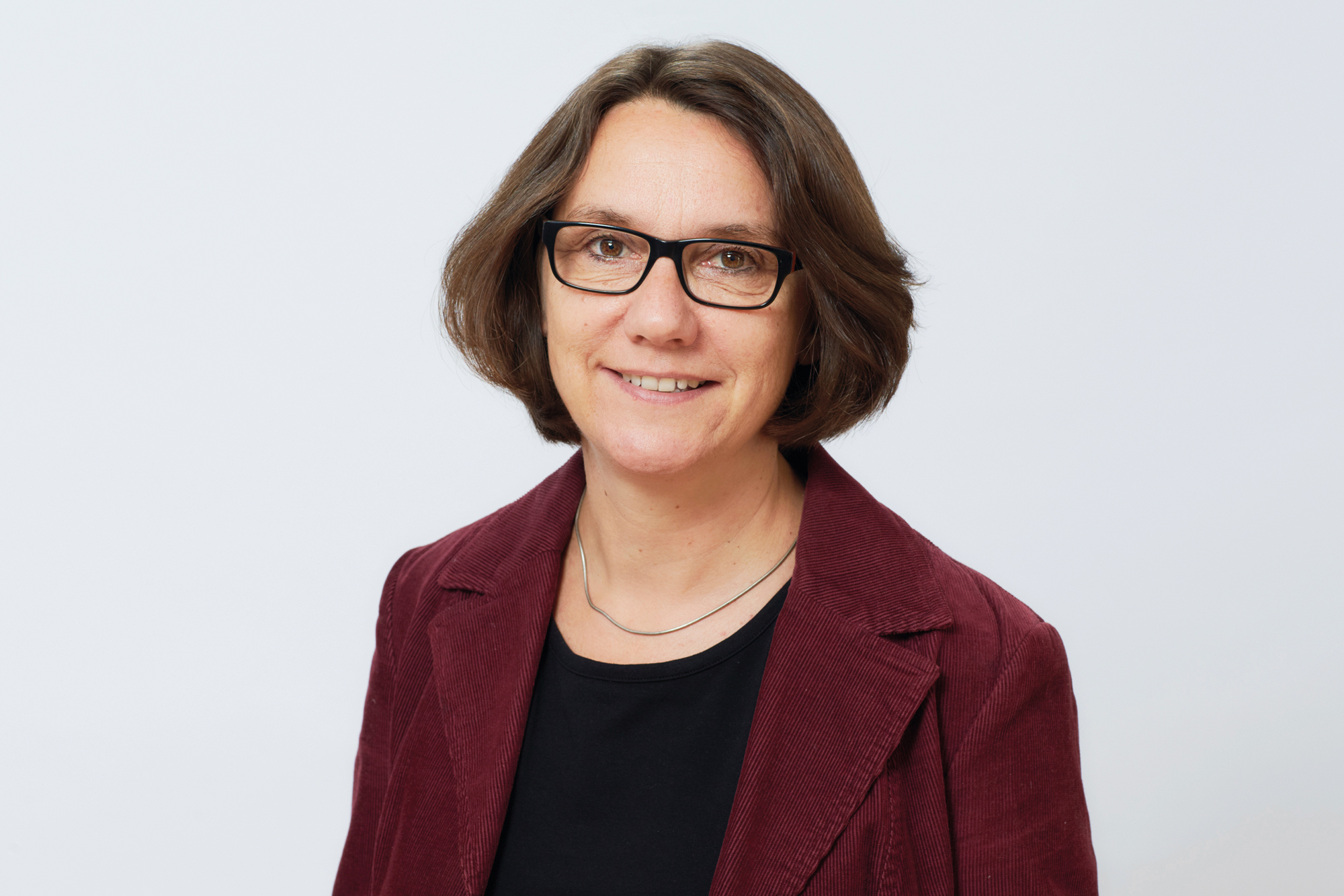 Eine Frau mit kurzen braunen Haaren und Brille lächelt in die Kamera und steht vor einem grauen Hintergrund.